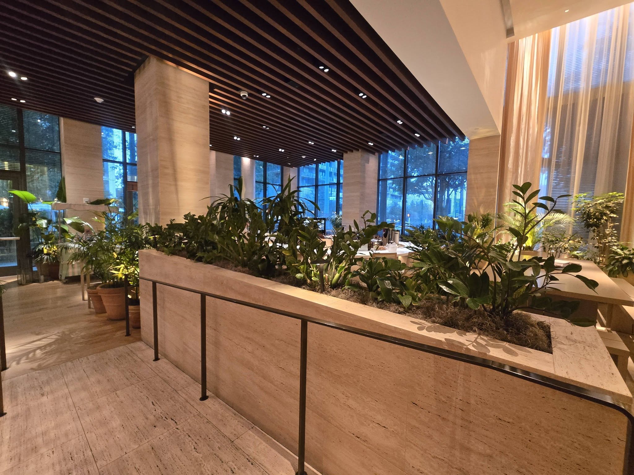 Long travertine built-in planter filled with ZZ plants, dracaena, and palms in a hotel lobby