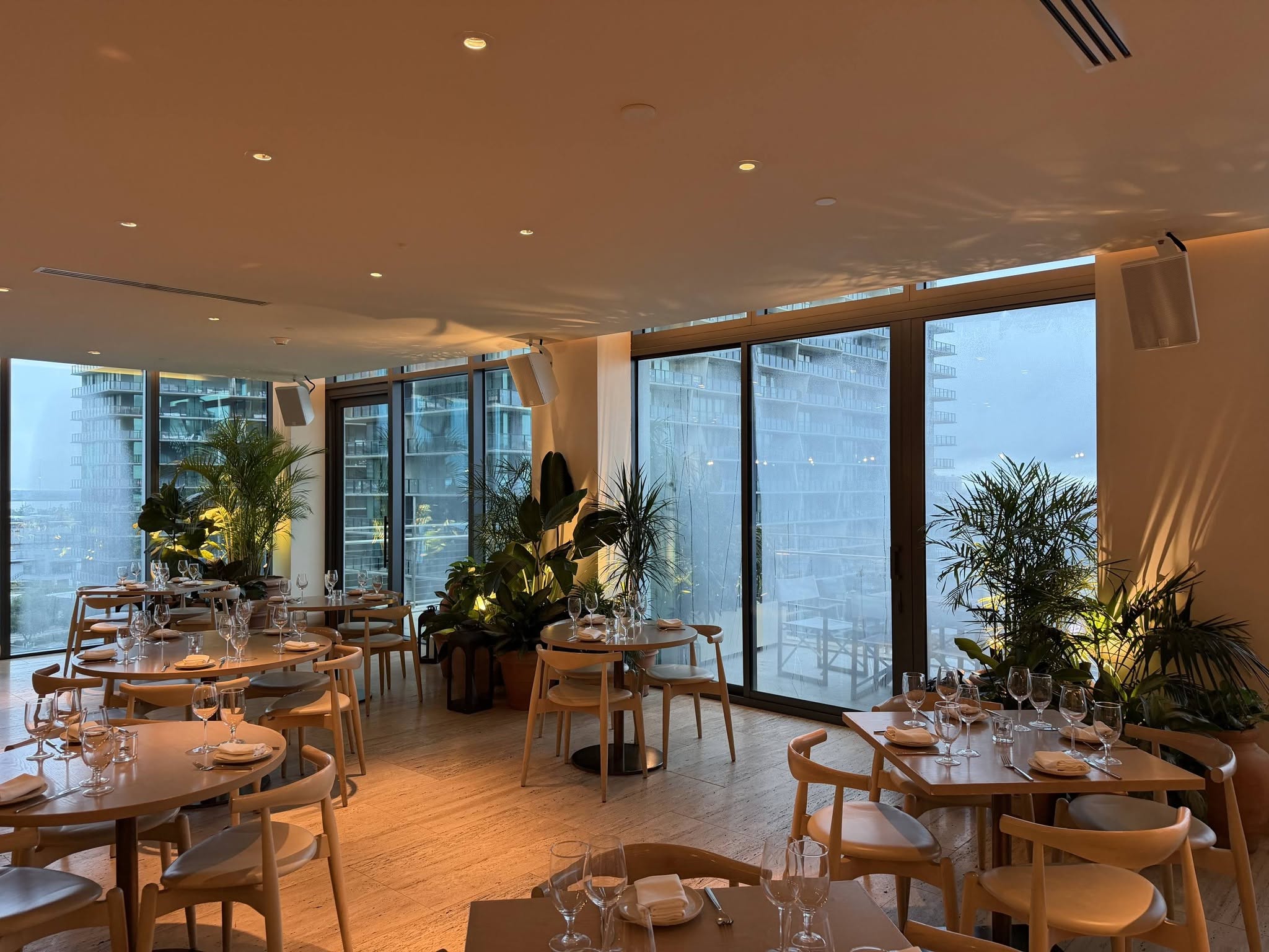 Hotel restaurant with round wooden tables and lush palm and bird of paradise groupings along the windows